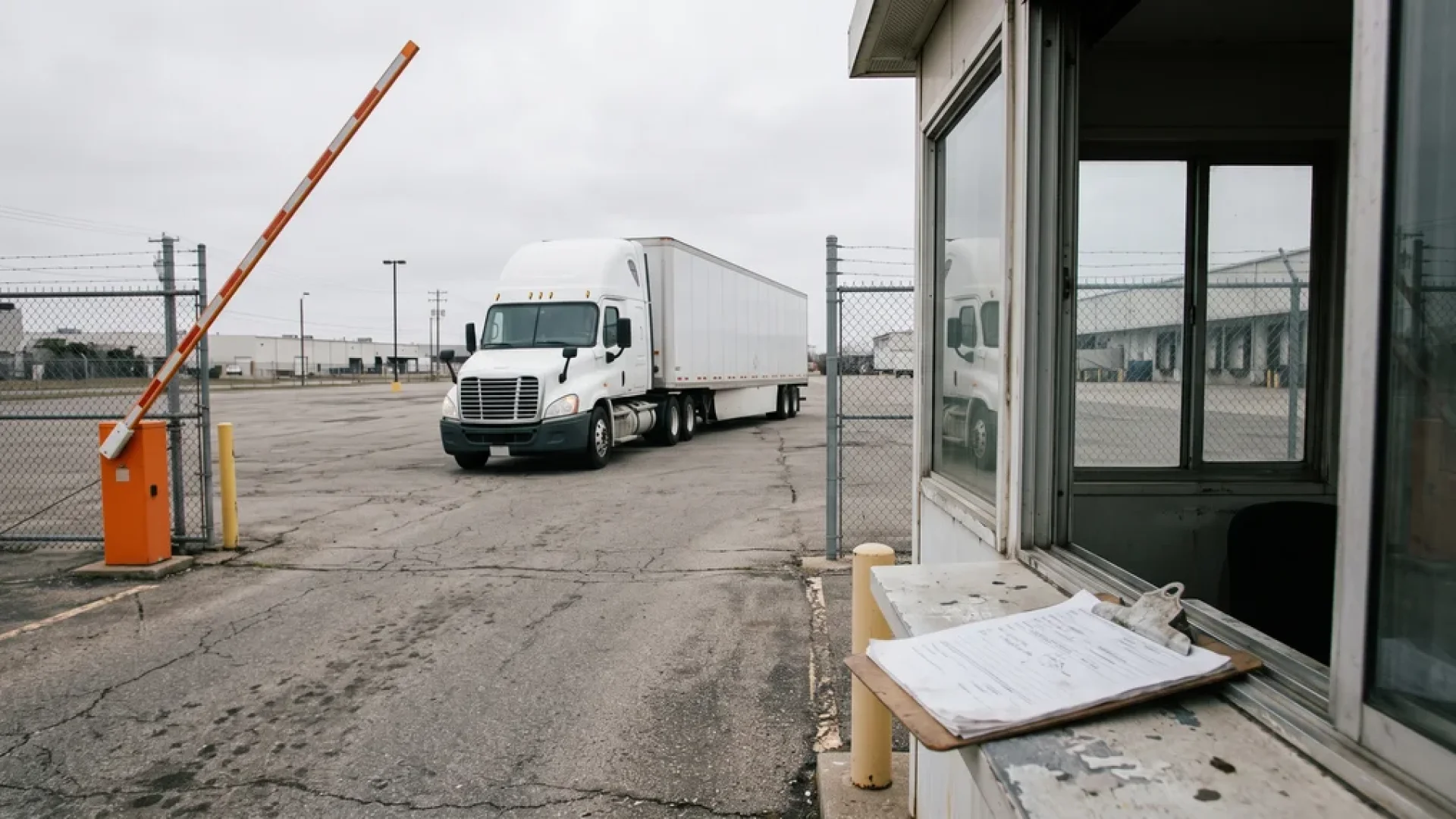Raised gate arm and empty guard booth at a warehouse yard as a white semi-truck pulls a trailer through the gate